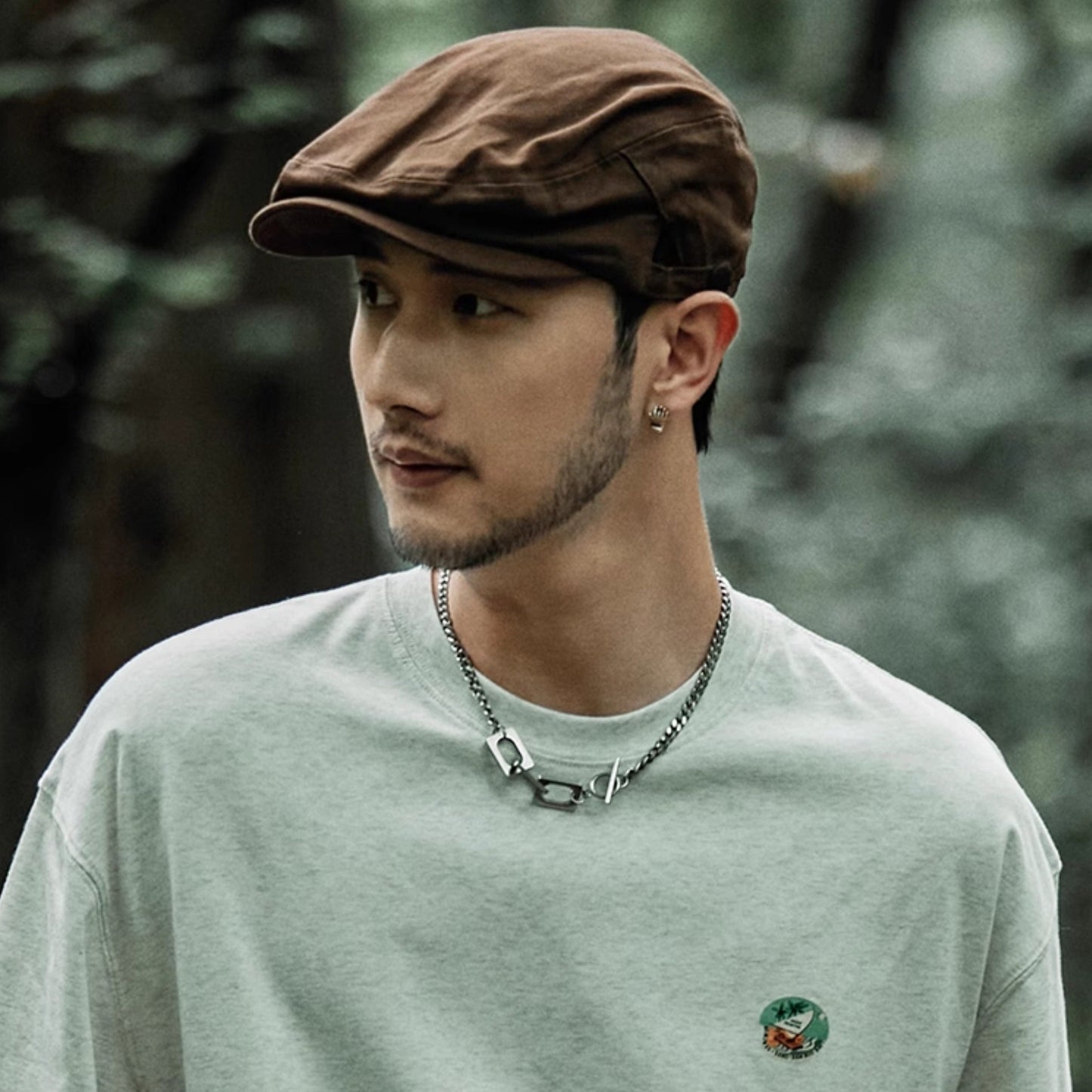 Man wearing a brown cap and light green shirt with a small logo, standing against a blurred natural background.