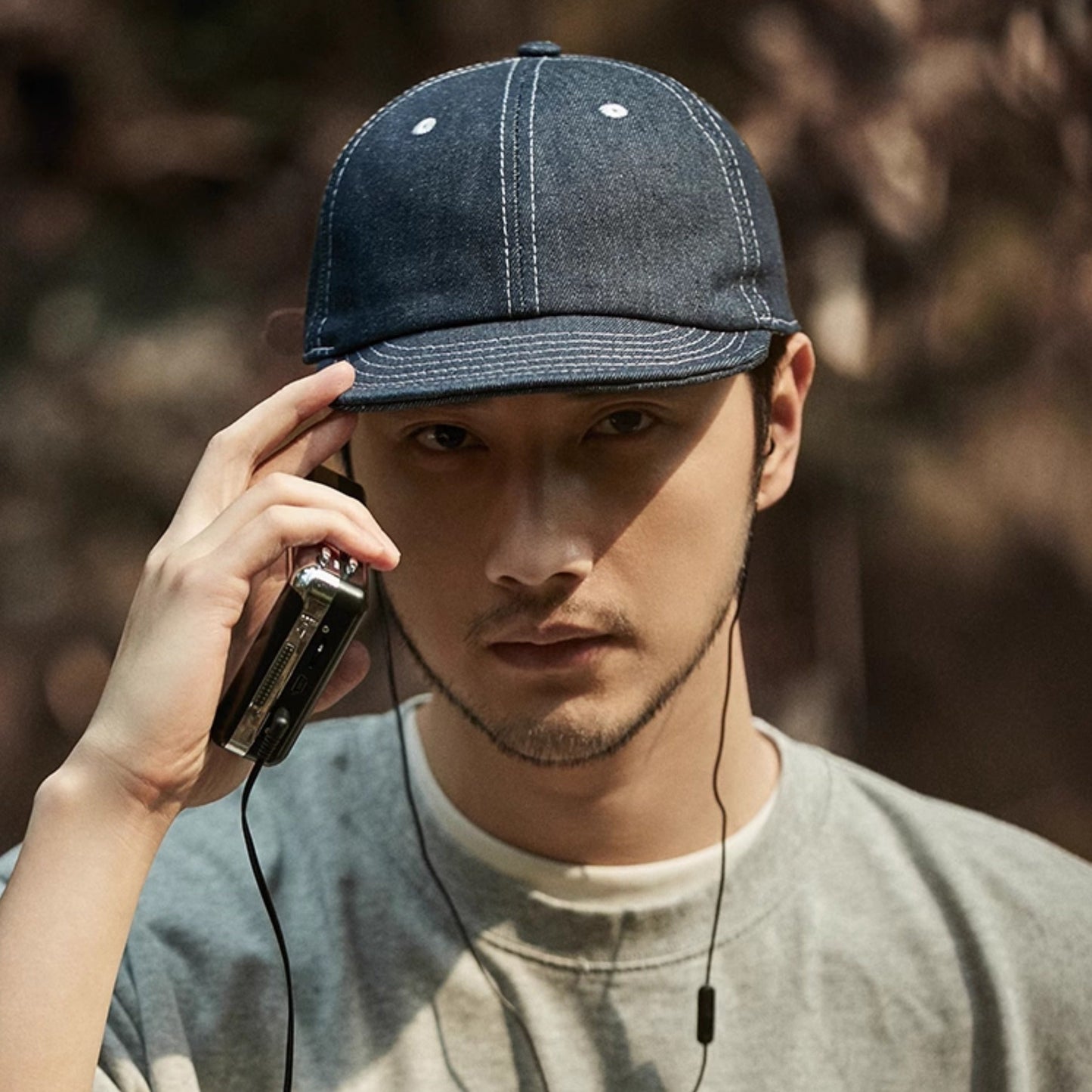 Man wearing a denim cap and listening to music with earphones, holding a phone to his ear.