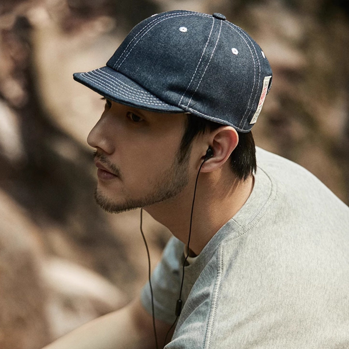 Man wearing a denim cap and listening to music with earphones, sitting outdoors.