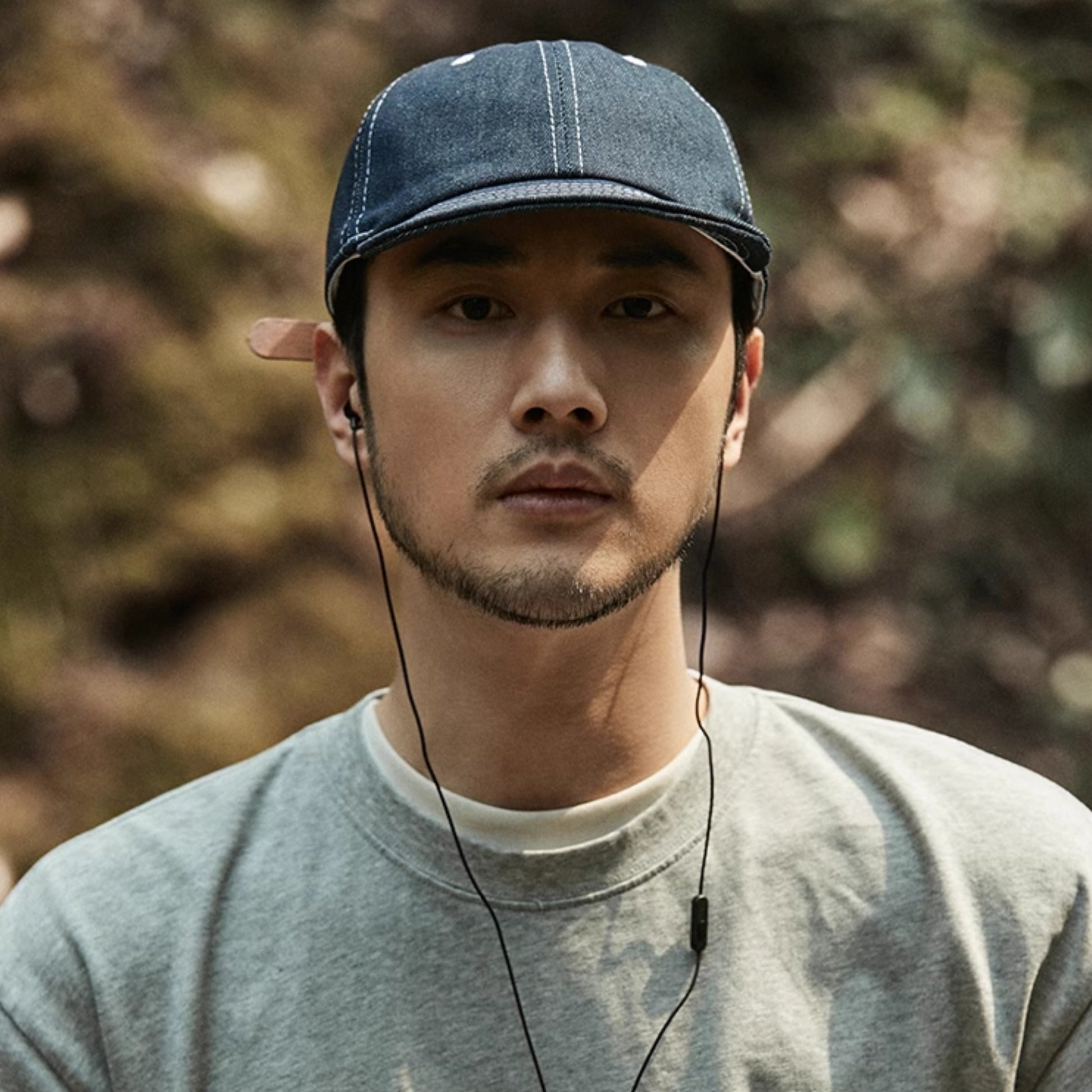 Man wearing a blue cap and gray sweatshirt with earphones, standing outdoors.