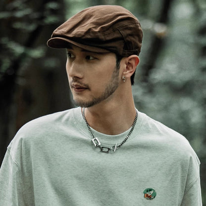 Man wearing a brown cap and light green shirt with a small logo, standing against a blurred natural background.